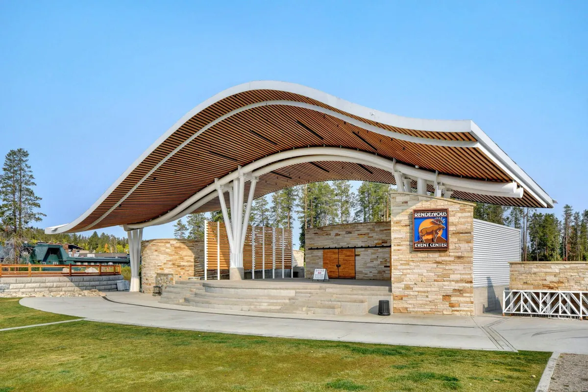 The Hideaway Park amphitheater with its iconic curved roof structure, stone walls, and open stage area. Signage and ample event space hint at concerts and community gatherings.