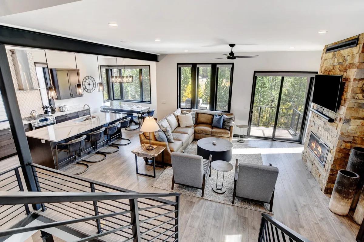 An open-plan living and kitchen area featuring a comfortable sectional sofa, a cozy fireplace, and a large kitchen island. The space seamlessly transitions to large windows offering views of the deck, inviting natural light and mountain scenery inside. The kitchen is integrated into the social hub, ideal for group meals and relaxed get-togethers.