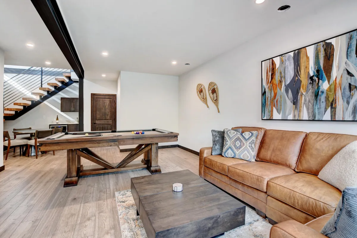 A stylish game room with a pool table, comfortable leather couch, modern wooden coffee table, and abstract wall art. The space is designed for friendly gatherings and socializing after a day outdoors.