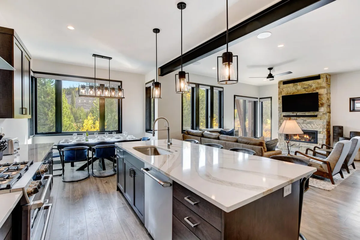 A spacious open-concept living area that seamlessly integrates a kitchen island, large floor-to-ceiling windows offering stunning mountain and river views, and a striking custom gas fireplace. Designer furnishings invite comfort, and the natural light fills the space, reinforcing the sense of connection with the outdoors.
