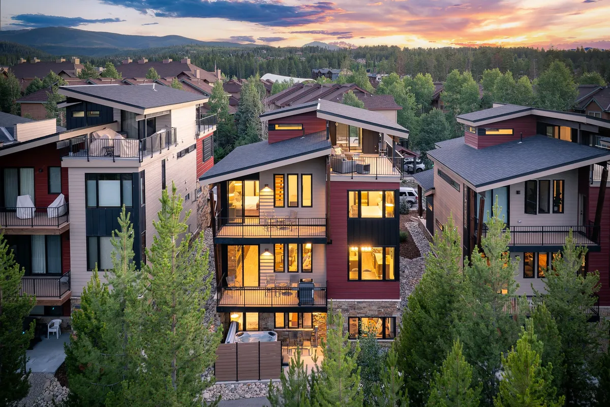 The image features a stunning modern chalet with expansive balconies, set against a breathtaking sunset sky. Guests are pictured relaxing on the balconies, enjoying panoramic views of the surrounding Colorado landscape. The scene evokes warmth and togetherness, with space for outdoor gatherings and a seamless connection to nature.