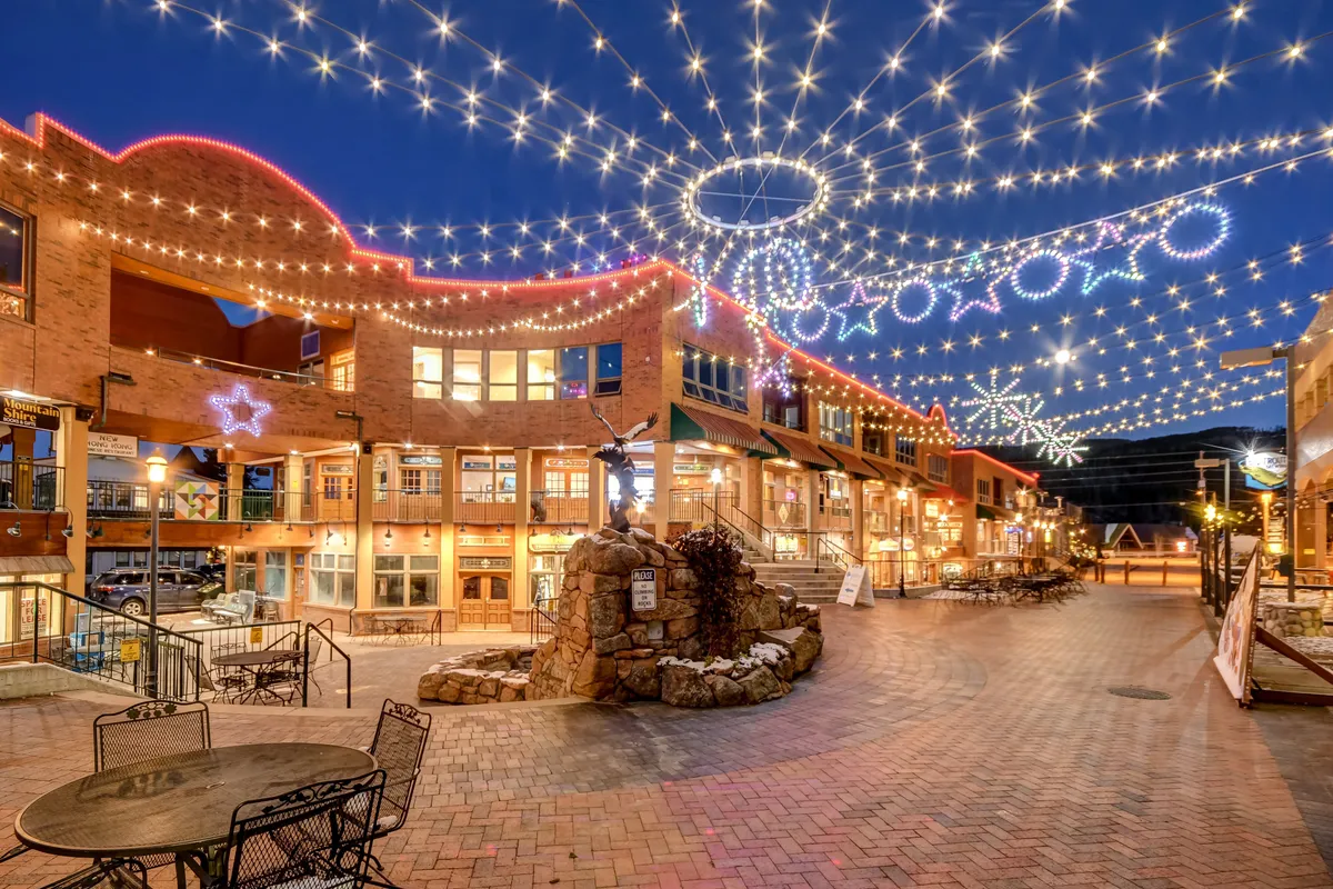 A charming downtown area illuminated by decorative string lights, featuring brick pathways, outdoor seating, and a fountain sculpture. People can be seen enjoying al fresco dining and evening strolls, evoking the lively festival atmosphere described for summer and the vibrant energy present year-round.