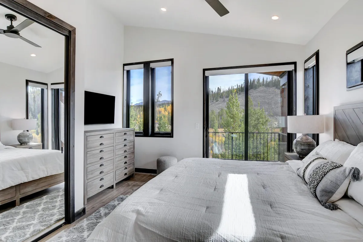 A spacious master bedroom featuring a king-sized bed with decorative pillows, large windows that flood the room with natural light and offer mountain views, a modern dresser with a television, and a mirrored wardrobe. The warm color palette and premium bedding evoke a sense of luxury and calm.