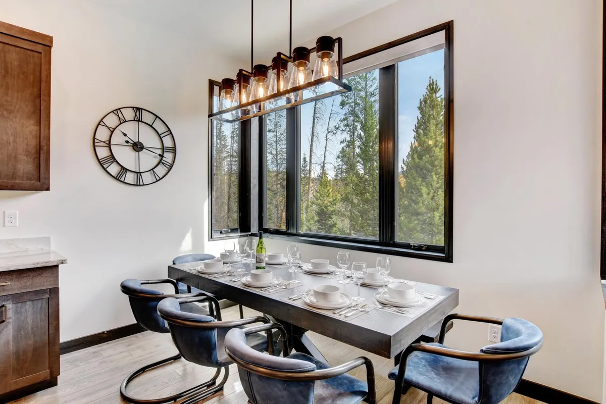 A warm, elegant dining area set for a meal, with large windows framing the natural landscape outside. The modern light fixture and decor evoke a sense of celebration and togetherness, perfect for shared meals and special occasions.