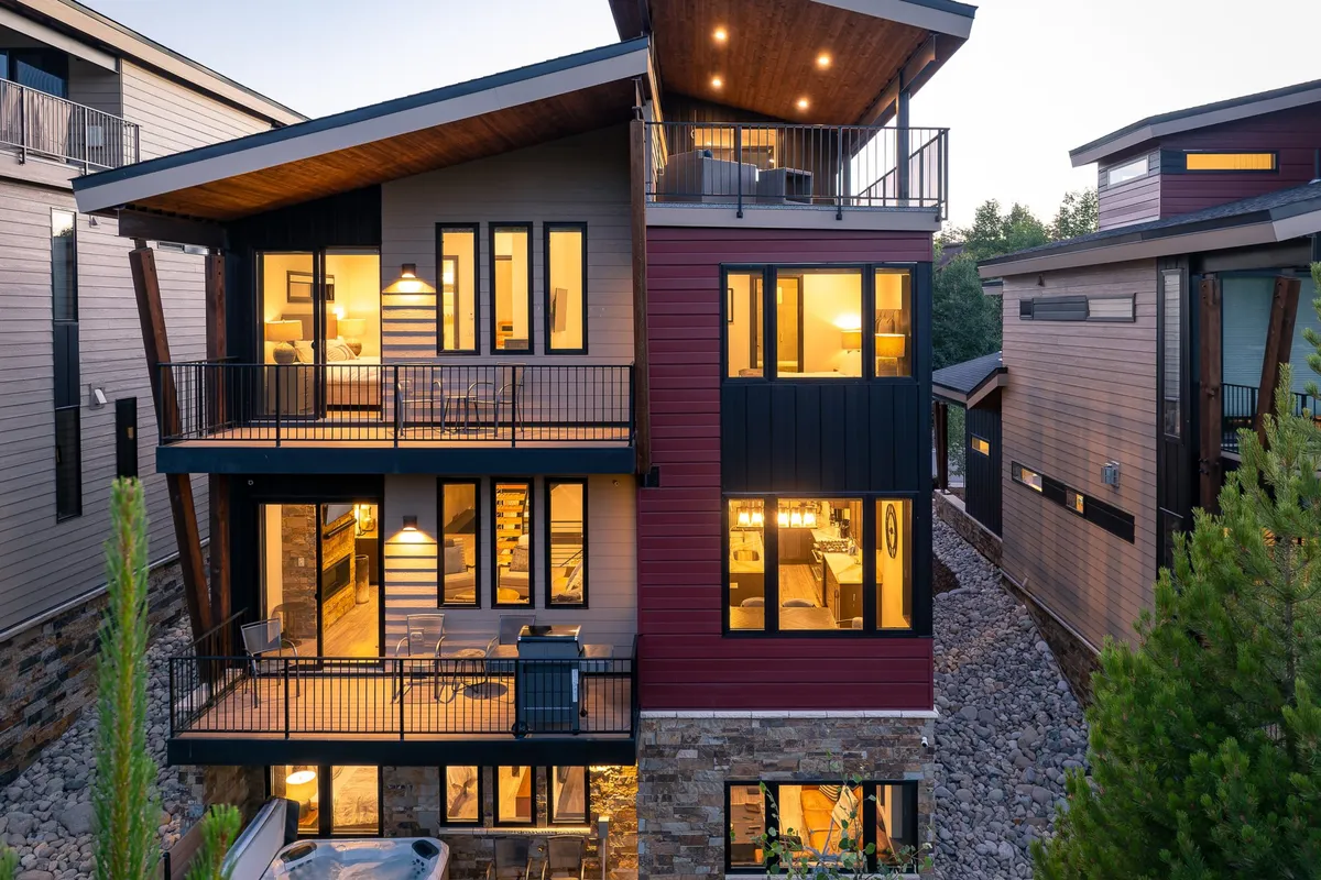An exterior view of a modern mountain home with expansive balconies, large windows, and an inviting outdoor hot tub area. The setting is framed by scenic mountain views and comfortable seating spaces for relaxation and gatherings.