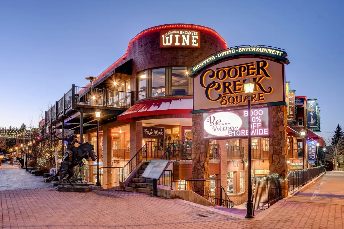 A vibrant outdoor plaza featuring the Cooper Creek Square sign, a wine shop entrance, outdoor seating, and a lively sculpture of a horse. Evening lights and people enjoying the space evoke a sense of community and social interaction.