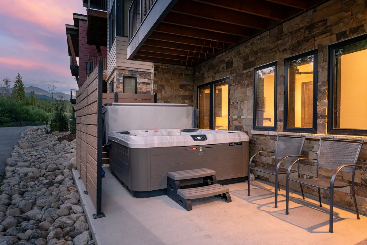 A tranquil outdoor space featuring a hot tub surrounded by stone elements and inviting outdoor chairs. The sky is aglow with sunset hues, suggesting a perfect moment for relaxation while taking in the mountain views.